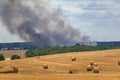 Harvested field with straw bales in summer on a sunny day with a field fire and smoke in the background