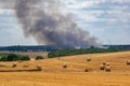 Harvested field with straw bales in summer on a sunny day with a field fire and smoke in the background