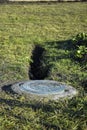 Hatch of well is closed with round metal cover. Sewer well with manhole. Trench is dug to well on green lawn.