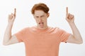 Hate rainy days. Portrait of moody displeased cute ginger guy with freckles in orange t-shirt, raising index fingers and