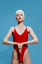 A haughty, serious young woman in a bathing cap and a red swimsuit folds her hands in meditation on a blue background