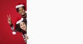 Smiling young african american male and female in santas hats peeks out from behind large white board Royalty Free Stock Photo