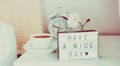 Have a nice day message on lighted box, alarm clock, cup of coffee and flower on the bedside table in sun light. Good morning mood Royalty Free Stock Photo