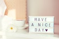 Have a nice day text message on lighted box, cup of coffee and white flower on the bedside table in sun light. Good morning mood. Royalty Free Stock Photo