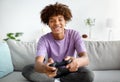 Having fun at home. Cheerful black teen guy with joystick playing online computer games, sitting on couch indoors