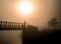 Hazy orange misty sunrise with lone man silhouette standing on river bridge looking at view of sun in fog. Stood wooden footbridge Royalty Free Stock Photo