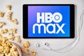 HBO MAX logo on the screen of the tablet laying on the white table and sprinkled popcorn on it. Apple earphones near the