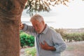 Head shot close up portrait sick old man feeling bad touching his chest at the park. Tired mature male resting next to tree