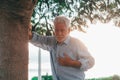 Head shot close up portrait sick old man feeling bad touching his chest at the park. Tired mature male resting next to tree