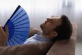 Head shot exhausted man waving paper fan, suffering from overheating