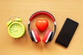 Headphones and alarm clock and smartphone and red heart on wooden desk. Musical concept