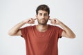 Headshot of annoyed stylish italian guy in red t shirt looking grumpy, closing ears with fingers showing disrespect on