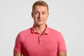 Headshot of attractive handsome man has appealing look, dressed in casual pink t shirt, ooks seriously at camera, over wh Royalty Free Stock Photo