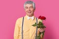 Headshot of attractive wrinkled senior male model looks happily at camera, dressed in elegant shirt, bowtie and suspenders, holds Royalty Free Stock Photo