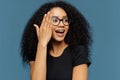 Headshot of beautiful curly woman looks aside, touches face, wears transparent glasses, black casual t shirt, isolated on blue