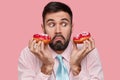 Headshot of handsome unshaven young guy looks with suprised facial expression, carries delicious doughnuts, frowns face