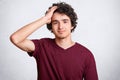 Headshot of handsome youngster, has curly hair, keeps hand on head, wears casual clothes, poses against white background. Joyful t