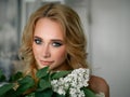 Headshot of happy beautiful blonde woman with lilac flower bouquet gorgeous trendy makeup beauty shiny skin posing indoors with