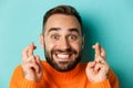 Headshot of hopeful bearded man making a wish, smiling and holding fingers crossed for good luck, standing over light