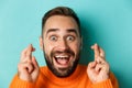 Headshot of hopeful bearded man making a wish, smiling and holding fingers crossed for good luck, standing over light