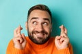 Headshot of hopeful bearded man making a wish, smiling and holding fingers crossed for good luck, standing over light