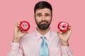 Headshot of puzzled unshaven man dressed formally, holds two doughnuts in both hands, going to eat pastry during lunch