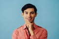 Headshot smiling handsome young adult latino man with hand on chin black hair and pink shirt over blue background