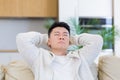 asian man holding his head with a severe headache at home in a room on the couch. Up close, the casual male