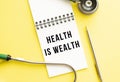 HEALTH IS WEALTH is written in a notebook on a color table next to pen and a stethoscope