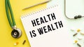 HEALTH IS WEALTH is written in a notebook on a color table next to pills and a stethoscope