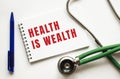HEALTH IS WEALTH is written in a notebook on a white table next to pen and a stethoscope.