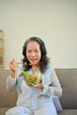 Healthy Asian 60s aged woman eating tasty Buddha salad vegetables mix bowl in living room Royalty Free Stock Photo