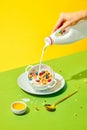 Healthy breakfast. Woman pouring milk into bowl with delicious muesli, cereal with berries against green yellow Royalty Free Stock Photo