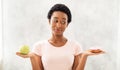 Healthy diet vs junk food. Black lady holding apple and donut in her hands, tempted to eat dessert instead of fruit Royalty Free Stock Photo