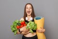 Healthy eating and active lifestyle. Amazed screaming young woman holding vegetables and yoga mat isolated on gray background Royalty Free Stock Photo