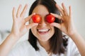 Healthy food concept. Young happy woman holding cherry tomato at eyes and smiling on background of modern white kitchen. Home Royalty Free Stock Photo