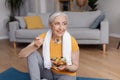 Healthy lifestyle concept. Happy senior woman eating fresh vegetable salad, sitting on yoga mat after home workout Royalty Free Stock Photo