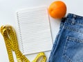 Healthy lifestyle and diet planning concept with orange, measuring tape, blank notebook, and denim jeans on white