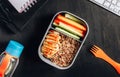 Healthy meal prep containers with quinoa, chicken and and sticks of cucumber and carrots on black wooden background. Healthy lunch
