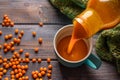 Healthy sea buckthorn juice is poured from a bottle into a blue mug on a wooden table. Berries and a warm scarf lie near