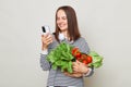 Healthy vegetarian diet. brown haired woman using smartphone for purchases holding fresh vegetables posing against gray background