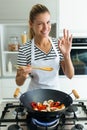 Healthy young woman looking at camera while cooking and mixing food in frying pan in the kitchen at home.