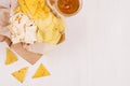 Heap different crispy golden snacks on craft paper, triangles nachos, lager beer in glass on soft white wood background.