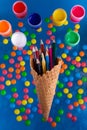 Heap of multicolor drawing pencils in ice cream waffle cone, colorful paints in round boxes on bright blue background with green,