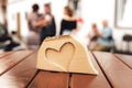 Heart shape carved in slice of wood laying on wooden table. Symbol of love. Blurred people in background.