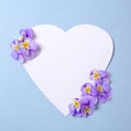 Heart-shaped blank paper card and wild violet flower petals on pastel blue background. Top view, floral frame, abstract design. Royalty Free Stock Photo