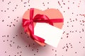 Heart shaped box with red ribbon bow and blank paper card mockup on pink background with confetti. Valentines day gift box. Love, Royalty Free Stock Photo