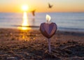 Heart shaped candle burning on sand of beach near sea waves during sunset dawn.