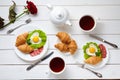 Heart shaped fried eggs, salad, croissants, salami sausage, rose flower composition and tea, white wooden table