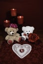 Heart shapped dollie and gemstone, three red candles in metal holoders and red rose, two teddy bears on wooden table.